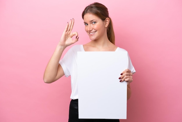 Joven mujer caucásica aislada sobre fondo rosa sosteniendo un cartel vacío y haciendo el signo de OK