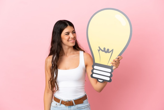 Joven mujer caucásica aislada sobre fondo rosa sosteniendo un icono de bombilla con expresión feliz