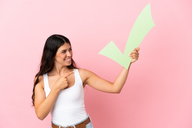 Joven mujer caucásica aislada sobre fondo rosa sosteniendo un icono de verificación y apuntando