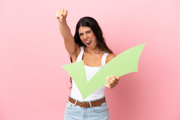 Joven mujer caucásica aislada sobre fondo rosa sosteniendo un icono de verificación y celebrando una victoria