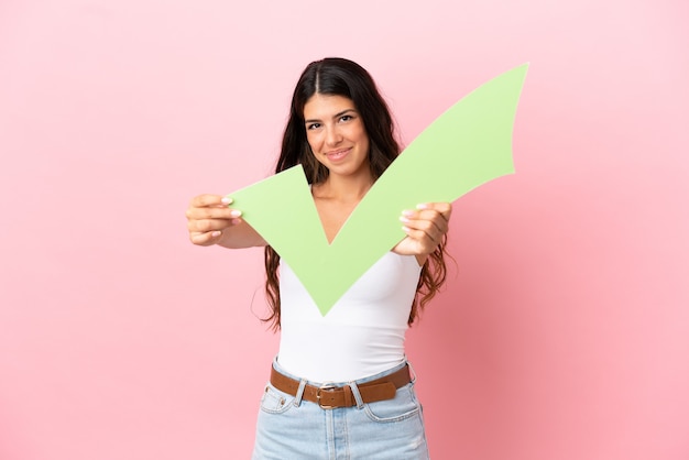 Joven mujer caucásica aislada sobre fondo rosa sosteniendo un icono de verificación con expresión feliz