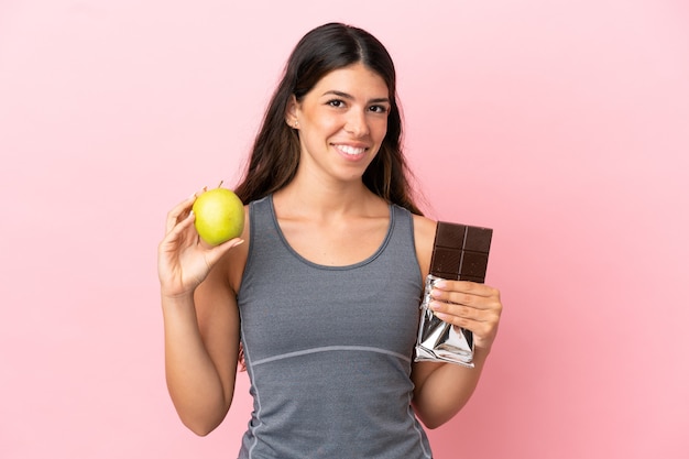 Joven mujer caucásica aislada sobre fondo rosa tomando una tableta de chocolate en una mano y una manzana en la otra
