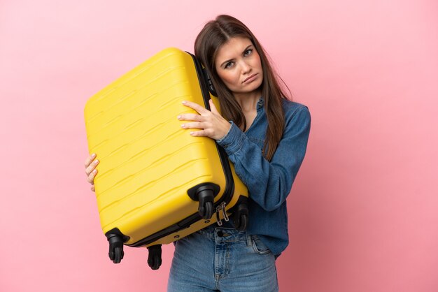 Joven mujer caucásica aislada sobre fondo rosa en vacaciones con maleta de viaje e infeliz