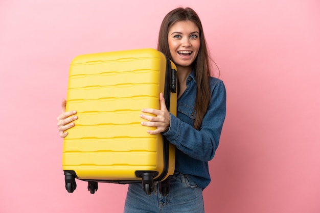 Joven mujer caucásica aislada sobre fondo rosa en vacaciones con maleta de viaje y sorprendido