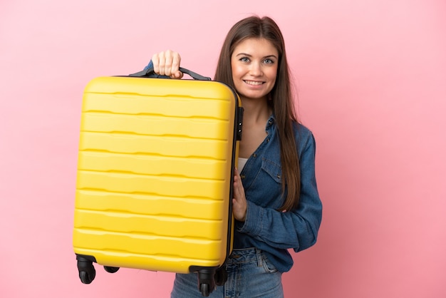 Joven mujer caucásica aislada sobre fondo rosa en vacaciones con maleta de viaje