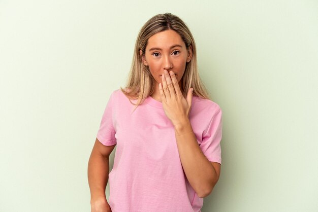 Joven mujer caucásica aislada sobre fondo verde conmocionada, tapándose la boca con las manos, ansiosa por descubrir algo nuevo.