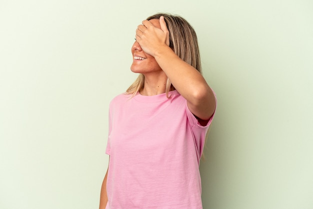 Joven mujer caucásica aislada sobre fondo verde cubre los ojos con las manos, sonríe ampliamente esperando una sorpresa.