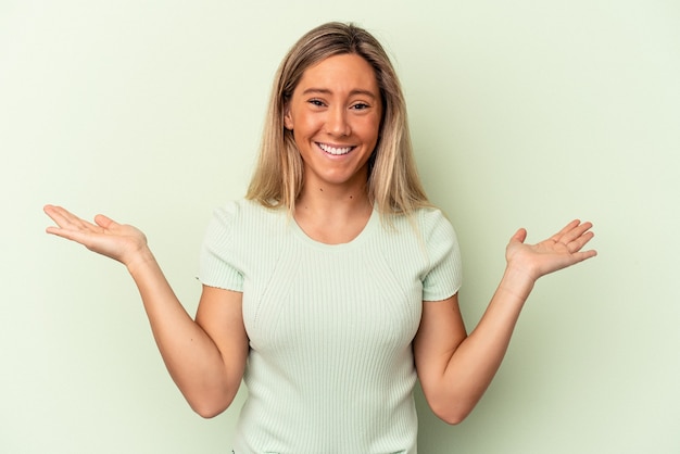 Joven mujer caucásica aislada sobre fondo verde hace escala con los brazos, se siente feliz y confiada.