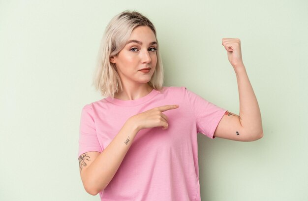 Joven mujer caucásica aislada sobre fondo verde mostrando gesto de fuerza con los brazos, símbolo del poder femenino