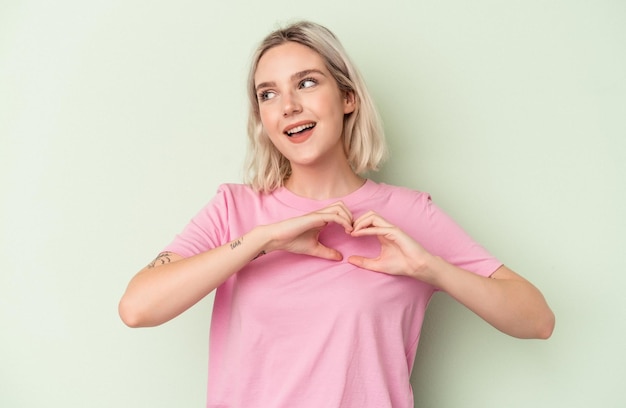 Joven mujer caucásica aislada sobre fondo verde sonriendo y mostrando una forma de corazón con las manos.