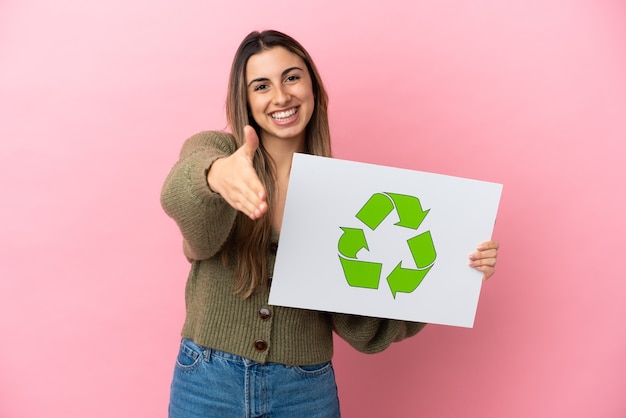 Joven mujer caucásica aislada sosteniendo un cartel con el icono de reciclaje haciendo un trato