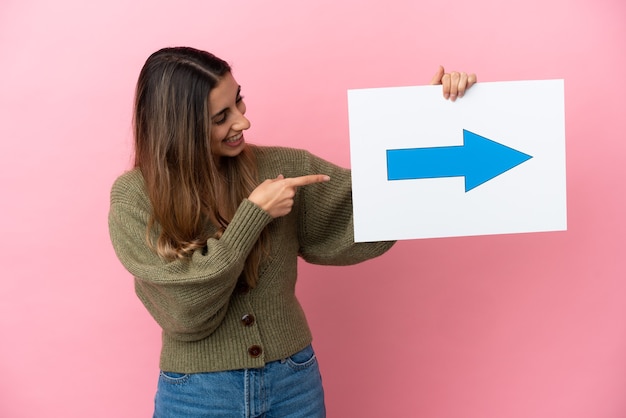 Joven mujer caucásica aislada sosteniendo un cartel con el símbolo de la flecha y apuntándolo