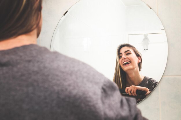 Joven mujer caucásica alisándose el cabello con un alisado de cabello en el espejo del baño.