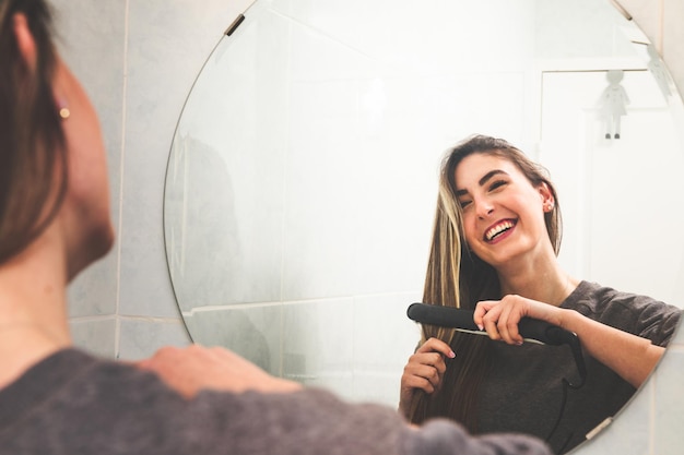 Joven mujer caucásica alisándose el cabello con un alisado de cabello en el espejo del baño.