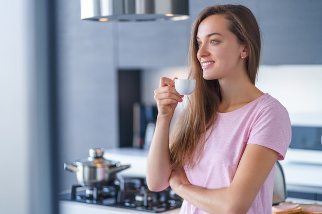 Joven mujer caucásica atractiva feliz bebiendo y disfrutando de café aromático fresco en casa en la cocina temprano en la mañana