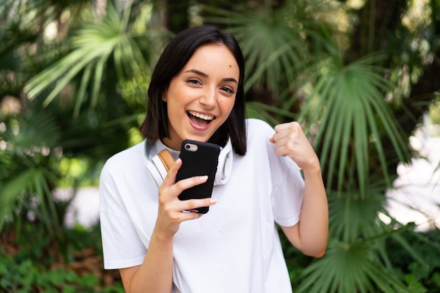 Joven mujer caucásica con auriculares al aire libre con teléfono en posición de victoria