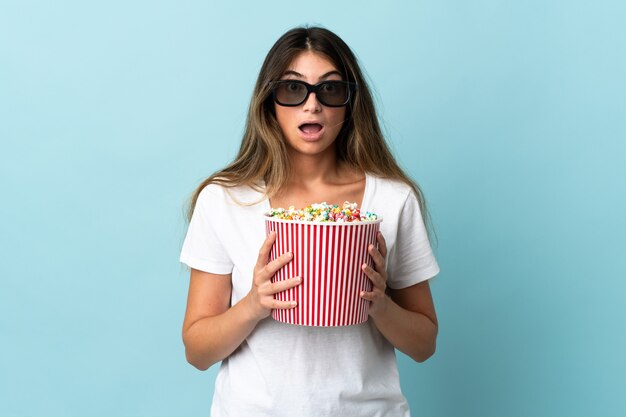 Joven mujer caucásica en azul sorprendido con gafas 3d y sosteniendo un gran cubo de palomitas de maíz