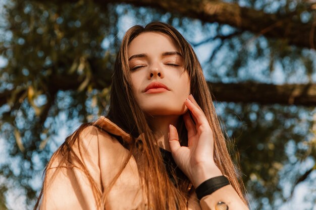 Joven mujer caucásica bastante elegante parado en el muelle de la calle cerca de un gran árbol vestido de trinchera marrón