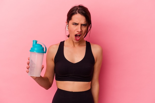 Joven mujer caucásica bebiendo un batido de proteínas aislado sobre fondo rosa gritando muy enojado y agresivo.