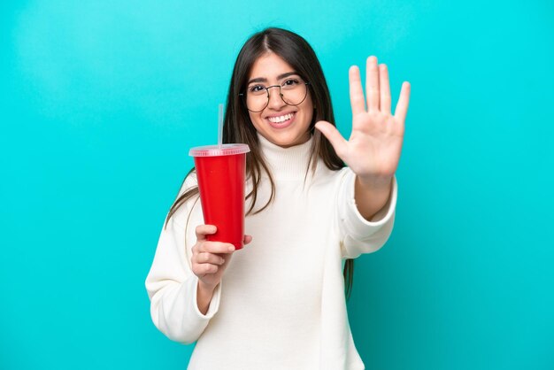 Joven mujer caucásica bebiendo soda aislada de fondo azul contando cinco con los dedos