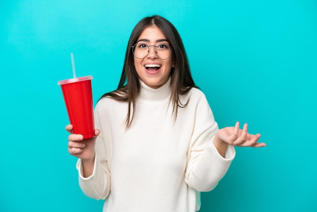 Joven mujer caucásica bebiendo soda aislada de fondo azul con expresión facial sorprendida