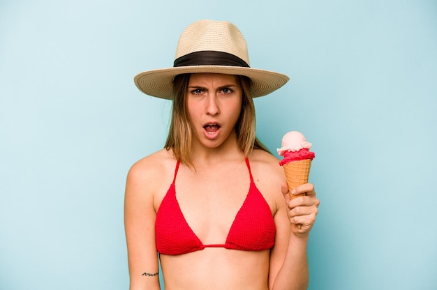 Joven mujer caucásica con bikini y sosteniendo un helado aislado de fondo azul gritando muy enojado y agresivo