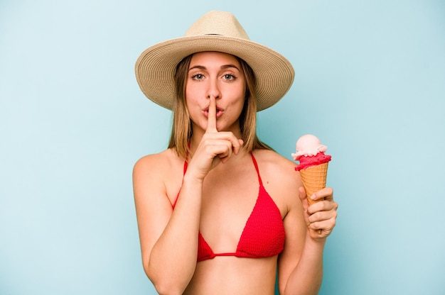 Joven mujer caucásica con bikini y sosteniendo un helado aislado de fondo azul guardando un secreto o pidiendo silencio