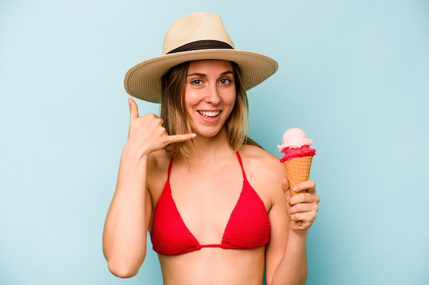 Joven mujer caucásica en bikini y sosteniendo un helado aislado de fondo azul mostrando un gesto de llamada de teléfono móvil con los dedos