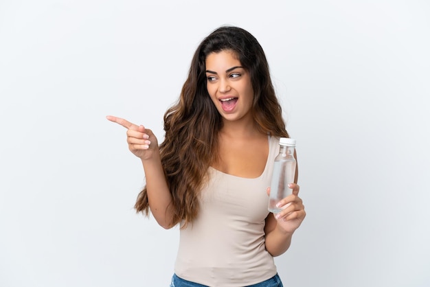 Joven mujer caucásica con una botella de agua aislado sobre fondo blanco apuntando con el dedo hacia un lado y presentando un producto
