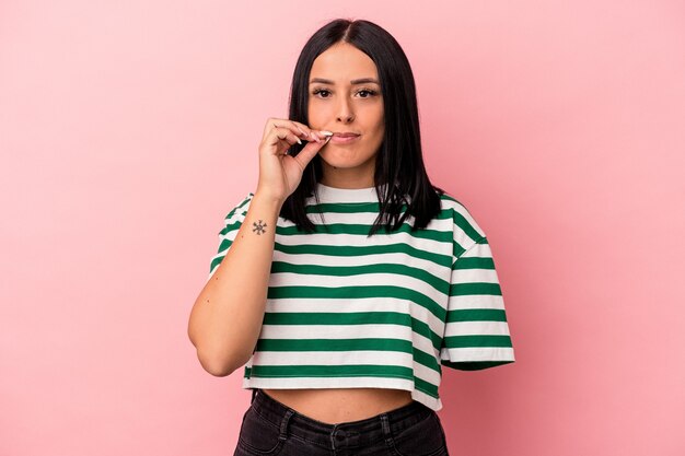 Joven mujer caucásica con un brazo aislado sobre fondo rosa con los dedos en los labios guardando un secreto.