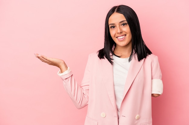 Joven mujer caucásica con un brazo aislado sobre fondo rosa mostrando un espacio de copia en una palma y sosteniendo otra mano en la cintura.