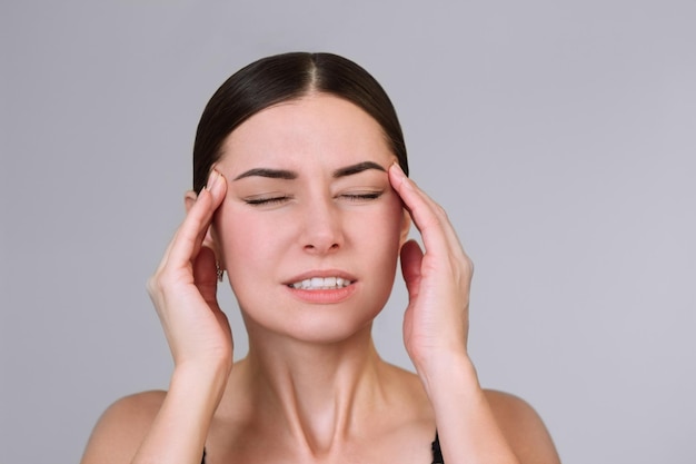 Una joven mujer caucásica de cabello oscuro con los ojos cerrados sosteniendo su cabeza con las manos doloridas