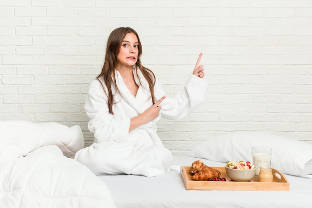 Joven mujer caucásica en la cama sorprendida señalando con los dedos índices a un espacio en blanco.