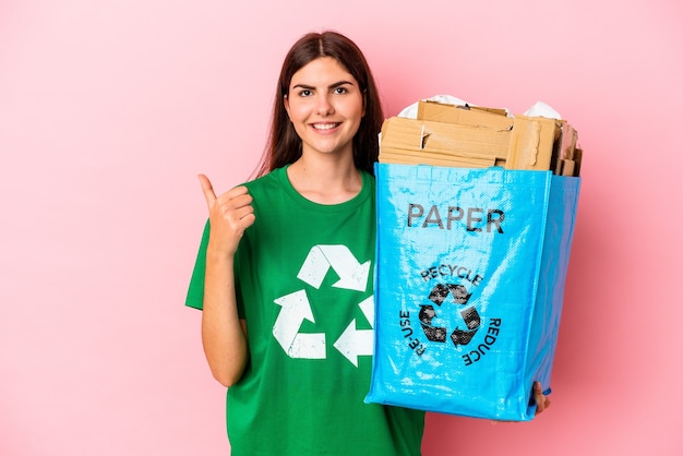 Joven mujer caucásica de cartón reciclado aislado en la pared rosa sonriendo y levantando el pulgar hacia arriba