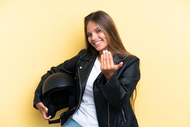 Joven mujer caucásica con un casco de motocicleta aislado en un fondo amarillo invitando a venir con la mano Feliz de que hayas venido