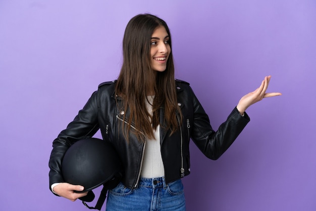 Joven mujer caucásica con un casco de motocicleta aislado sobre fondo púrpura extendiendo las manos hacia el lado para invitar a venir