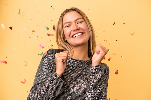 Joven mujer caucásica celebrando el año nuevo con confeti aislado sobre fondo amarillo