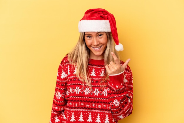 Joven mujer caucásica celebrando la Navidad aislado sobre fondo amarillo apuntando con el dedo hacia usted como si estuviera invitando a acercarse.