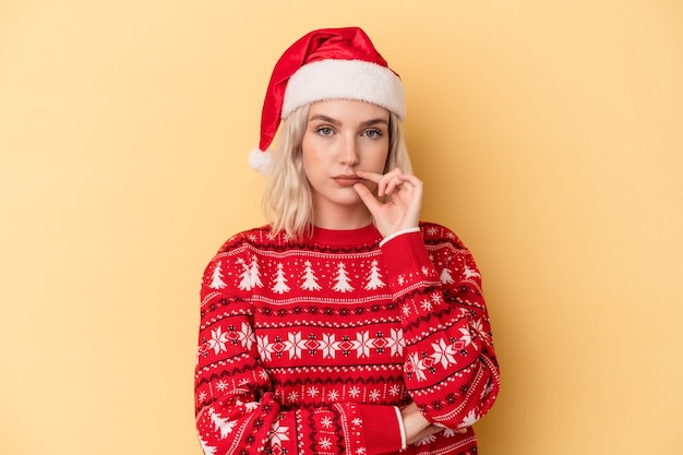 Joven mujer caucásica celebrando la Navidad aislado sobre fondo amarillo con los dedos en los labios guardando un secreto.