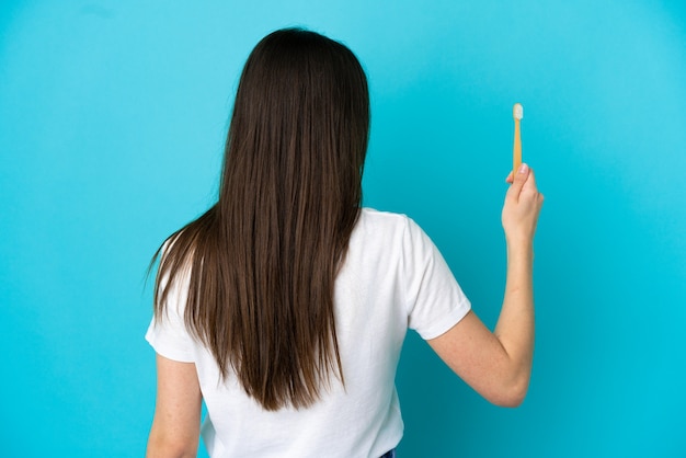 Joven mujer caucásica cepillarse los dientes aislado sobre fondo azul en posición trasera