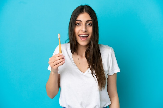 Joven mujer caucásica cepillarse los dientes aislado sobre fondo azul con sorpresa y expresión facial conmocionada