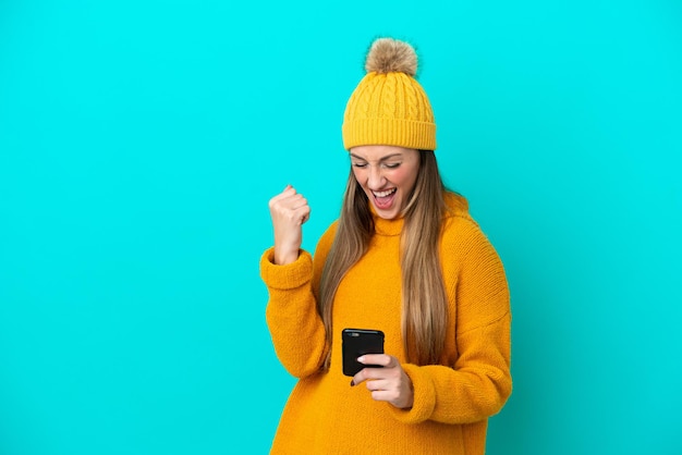 Joven mujer caucásica con chaqueta de invierno aislada de fondo azul con teléfono en posición de victoria