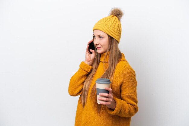 Joven mujer caucásica con chaqueta de invierno aislada de fondo blanco sosteniendo café para llevar y un móvil