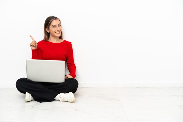 Joven mujer caucásica con una computadora portátil sentada en el suelo mostrando y levantando un dedo en señal de lo mejor