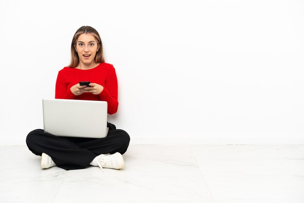 Joven mujer caucásica con una computadora portátil sentada en el suelo sorprendida y enviando un mensaje