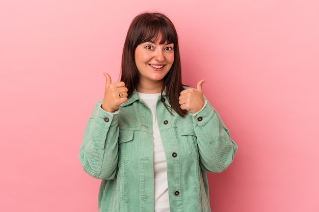 Joven mujer caucásica con curvas aislada sobre fondo rosa levantando ambos pulgares, sonriendo y confiado.