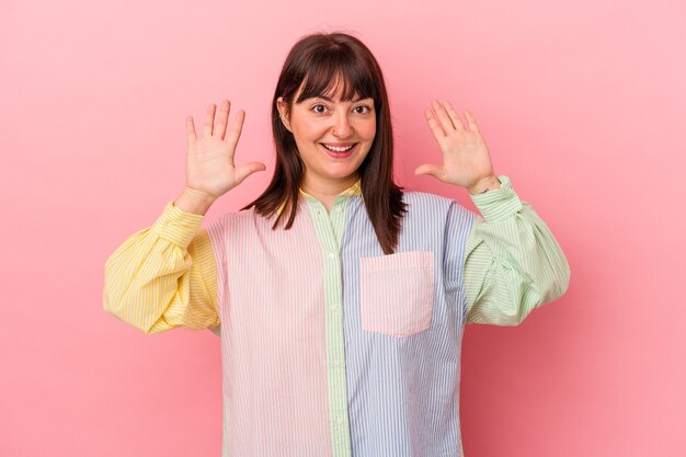 Joven mujer caucásica con curvas aislada sobre fondo rosa mostrando el número diez con las manos.