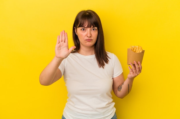 Joven mujer caucásica con curvas sosteniendo papas fritas aisladas sobre fondo amarillo de pie con la mano extendida mostrando la señal de stop, previniéndote.