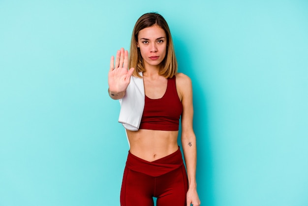 Joven mujer caucásica de deporte aislada en la pared azul de pie con la mano extendida mostrando la señal de stop, impidiéndote.