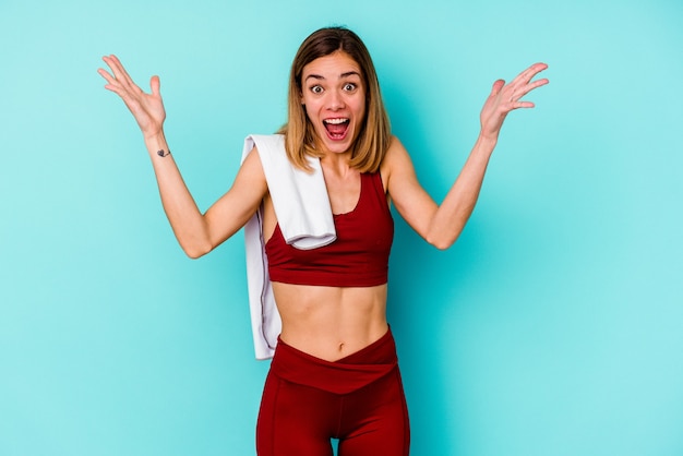 Joven mujer caucásica de deporte aislada en la pared azul recibiendo una agradable sorpresa, emocionada y levantando las manos.